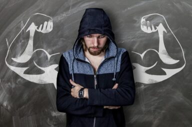 Image of a man in a hoodie standing in front of a drawing of muscular flexed arms on the National Personal Training Institute website