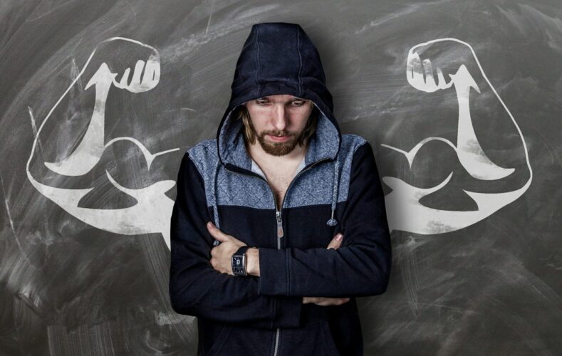 Image of a man in a hoodie standing in front of a drawing of muscular flexed arms on the National Personal Training Institute website