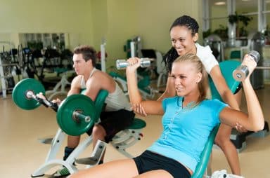 Young woman lifting a dumb-bell in the gym assisted by her personal trainer
