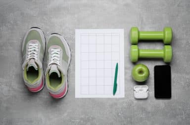 Image of a pair of sneakers, two hand weights, an apple, a set of headphones, a phone, a pen, and a planner on a gray floor