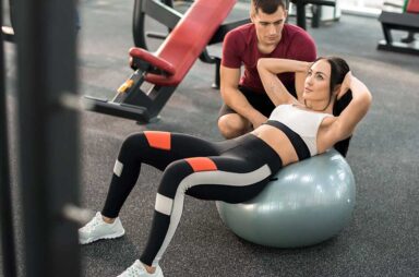 High angle portrait of muscular coach helping young woman doing exercises on fitness ball in modern gym