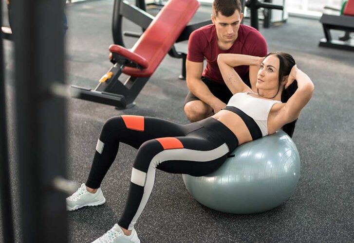 High angle portrait of muscular coach helping young woman doing exercises on fitness ball in modern gym