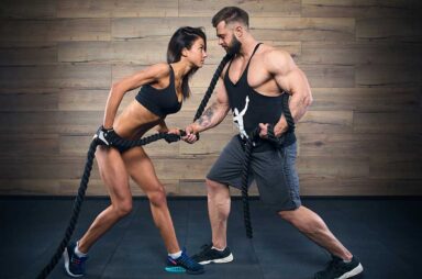 A sporty girl and a muscular shredded man with a beard are pulling a rope during a CrossFit training in a gym. A bodybuilder with a tattoo is training with a fitness girl in black tank tops and shorts