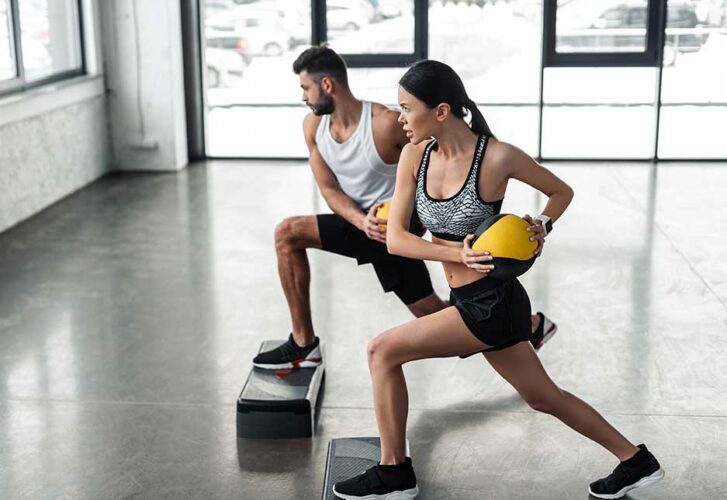 Side view of sportive young couple holding medicine balls and exercising on step platforms in gym