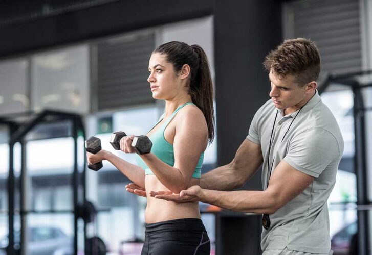 Male trainer assisting woman lifting dumbbells